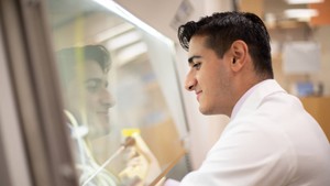A close up image of a male student in a lab coat working in a lab with his reflection visible in the glass.