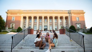 Four female students talking and laughing while seated on the steps of Dinand Library.