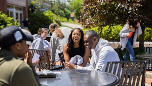 Two students laugh while seated at an outdoor table while other students interact around them.