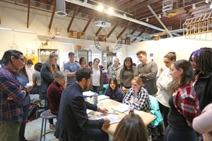 A large group of students clustered around a professor who is  seated at a table in an art studio setting.