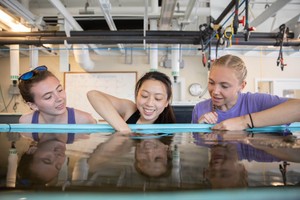 Three female students smiling and working in a laboratory in front of a tank of water with one student reaching into the water.