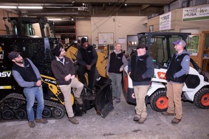 A group of six Holy Cross facilities workers smile and interact while standing around heavy equipment in a garage setting.