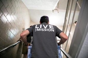 The back of a male student carrying luggage up a staircase wearing a shirt that reads “Live The Mission.”