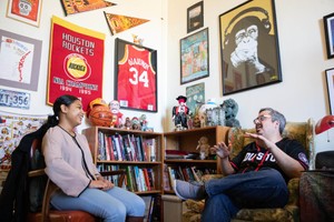 A female student talks with a teacher while seated in his office which is heavily decorated with sports memorabilia.