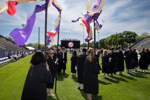 Four flag bearers wearing graduation gowns in the back of a line of students carry colorful flags that flutter in the wind towards the camera.