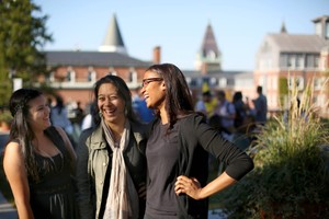 Three female students smiling and interacting with each other with campus buildings visible in the background.