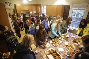 A large group of students fills a kitchen in the foreground of which is a table filled with bagels.