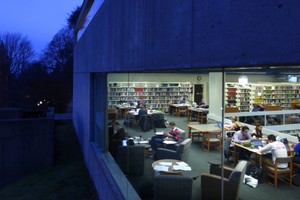 A view at dusk looking inside a large library window at students seated around desks while studying.
