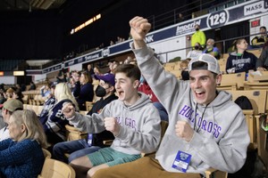 Two male students wearing Holy Cross sweatshirts cheer at a hockey game at the DCU Center in Worcester, MA.