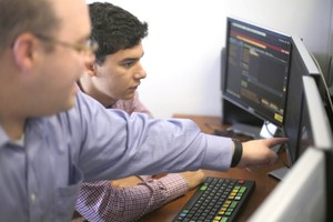 A professor points to a screen while working with a student at a work station containing four monitors.